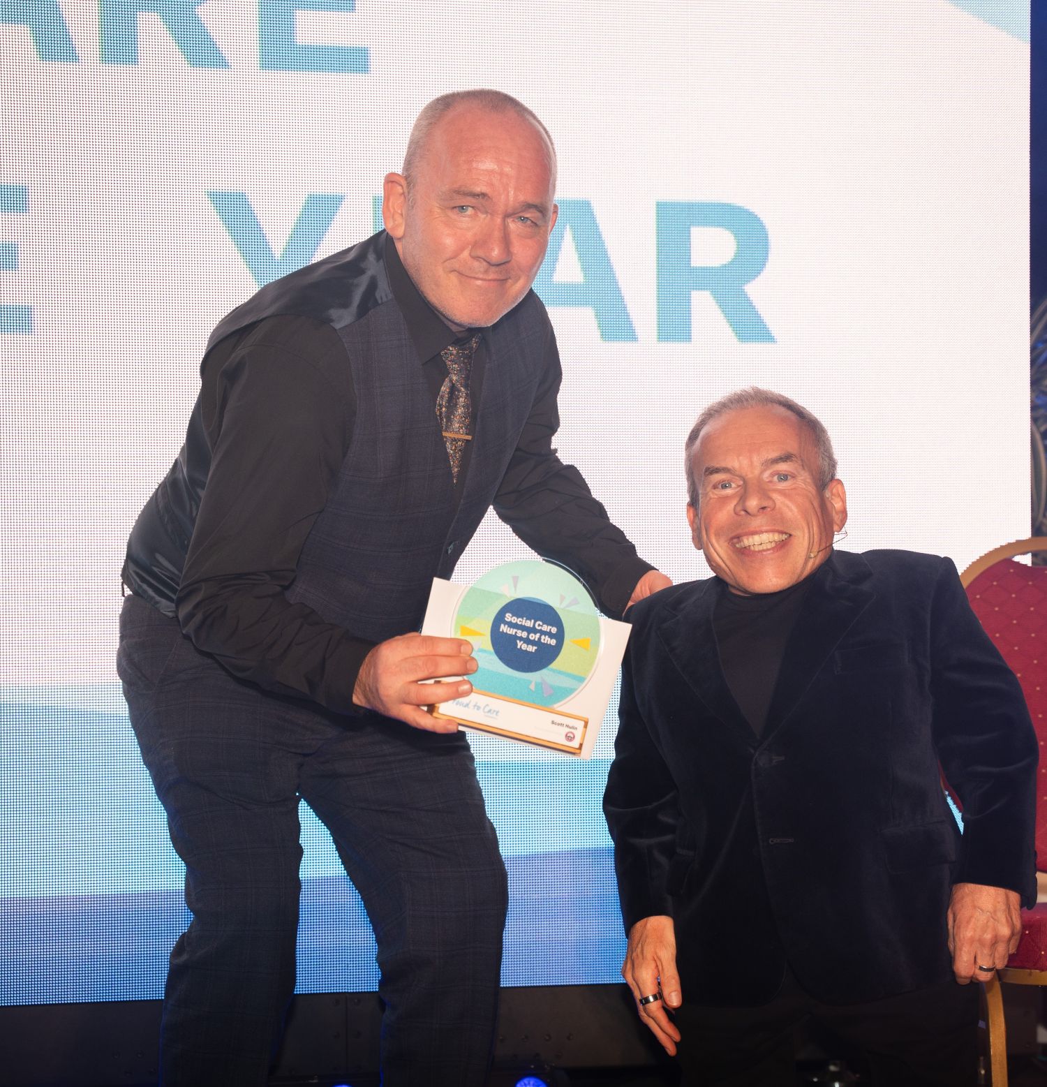A man recieving a care award from the actor Warwick Davis